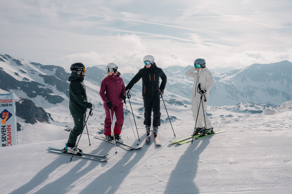 4 Skifahrer stehen auf der Piste, im Hintergrund ist die schneebedeckte Berglandschaft zu sehen