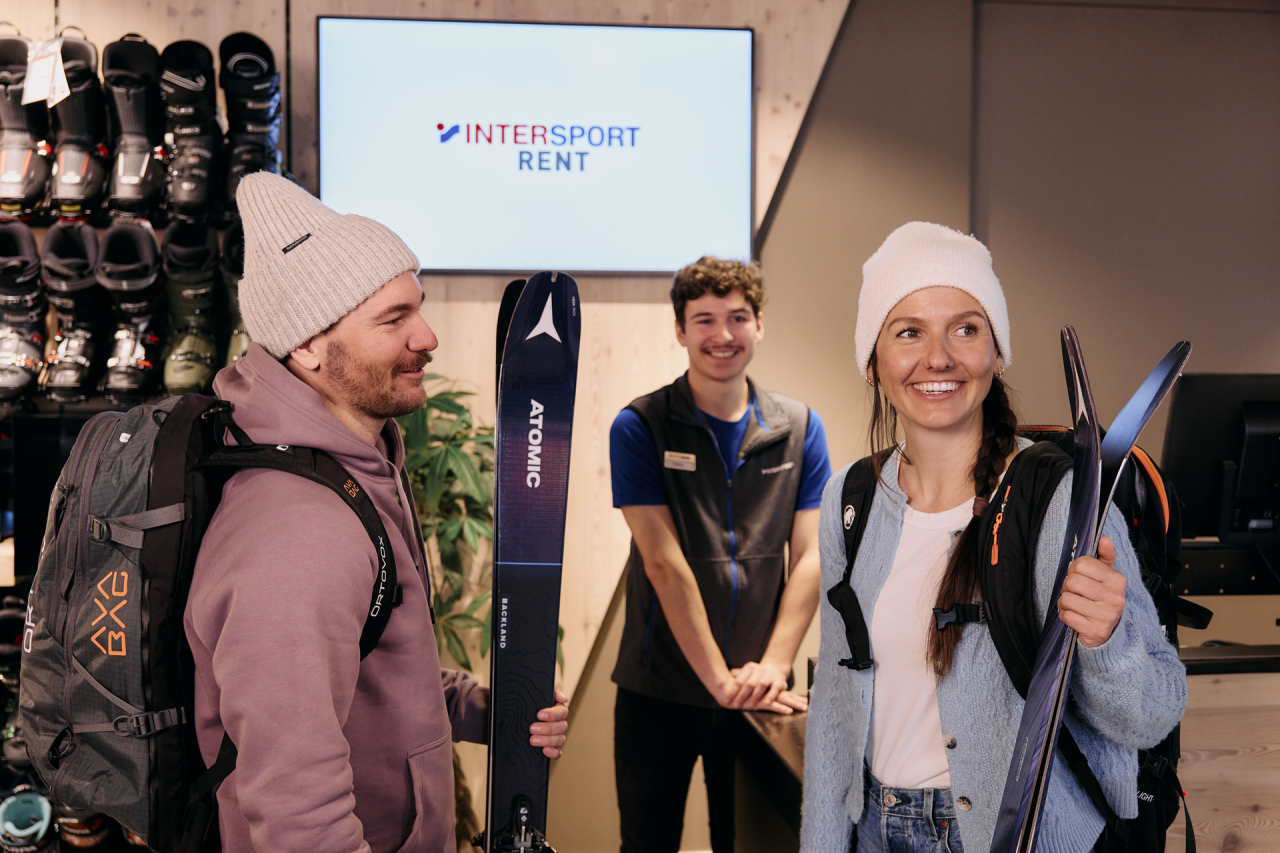 Two customers are standing in the shop holding skis in their hands, with an employee standing in the background