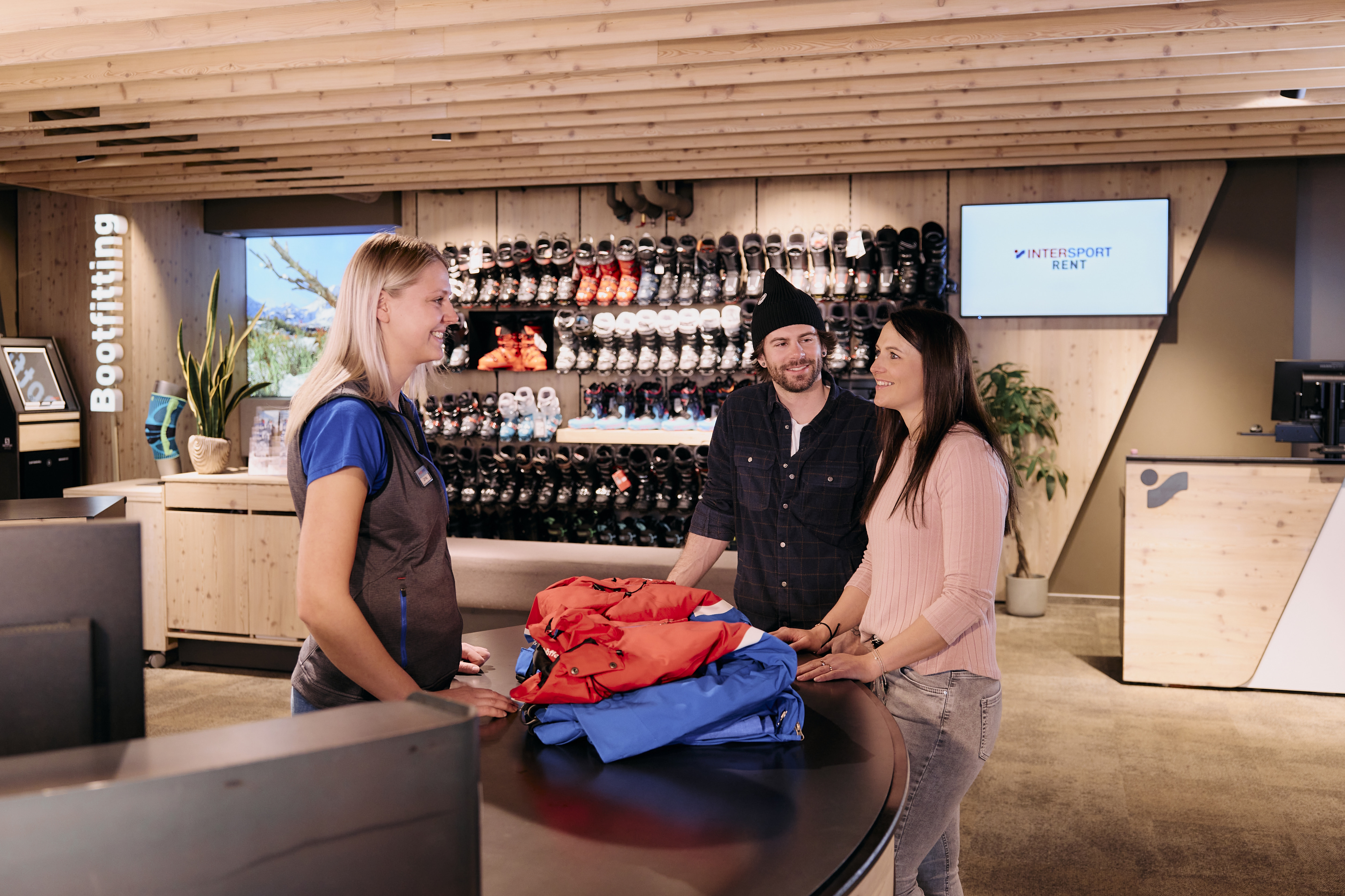 Zwei Kunden im Sportgeschäft im Gespräch mit einer Mitarbeiterin von INTERSPORT an der Servicetheke, im Hintergrund eine Wand mit Skischuhen.