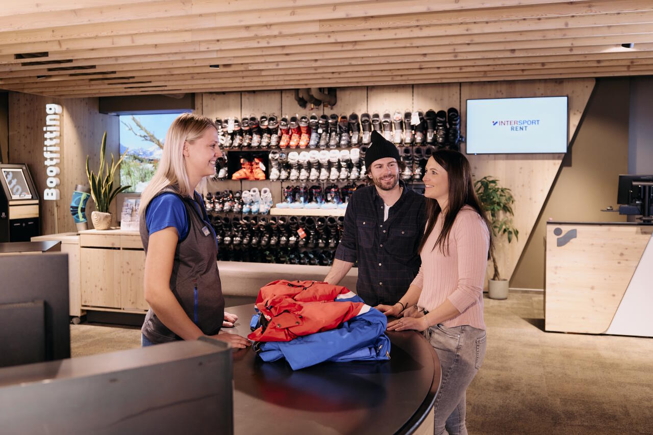Two customers in a sports shop talking to an INTERSPORT employee at the service counter, with a wall of ski boots in the background.